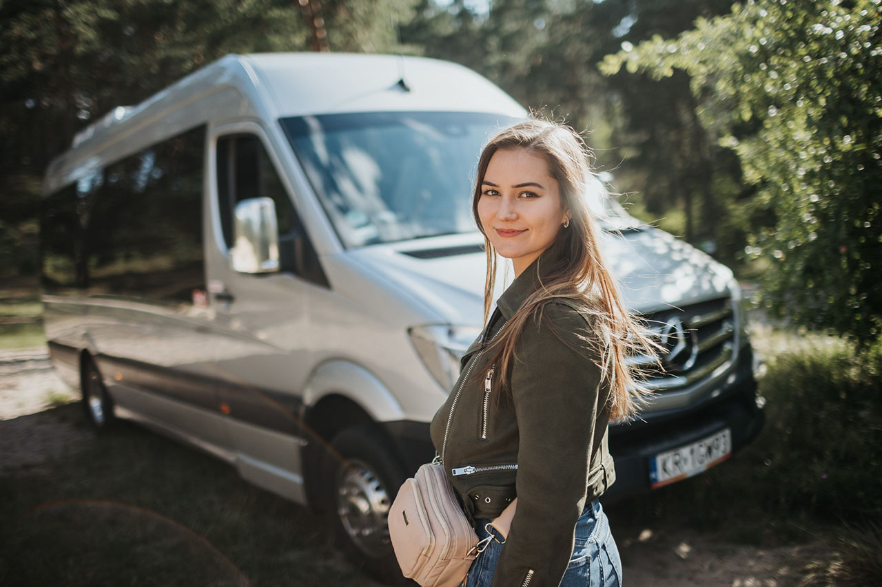 Smiling woman standing next to mini bus surrounded by green nature